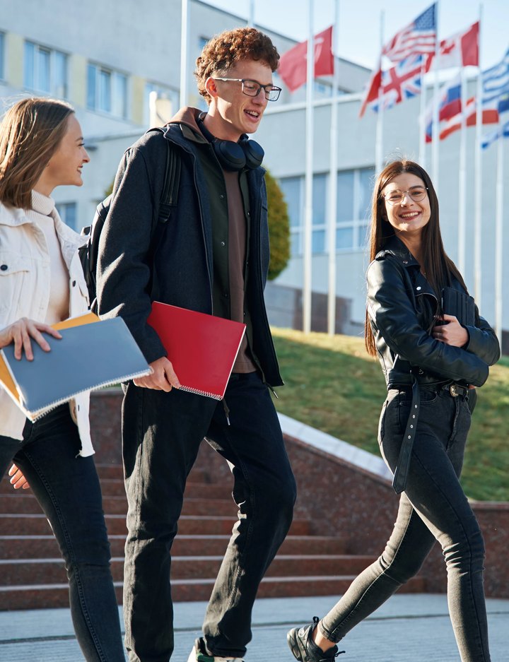 WMK — three students with notebooks walking past a building with flags